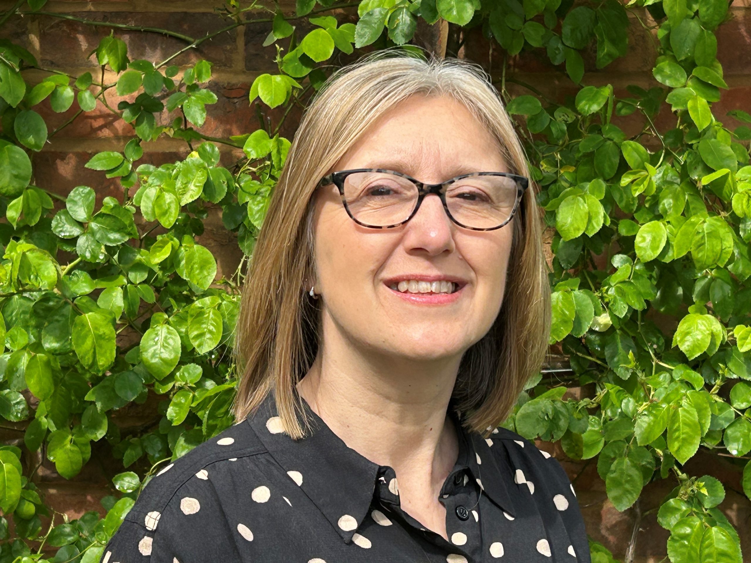 An image of Carolyn Tarrant, with shoulder-length straight hair and glasses, standing in front of greenery